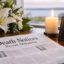 Newspaper displaying recent death notices and obituaries beside white lilies, a glowing candle, and a memorial card.
