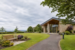 A modern crematorium surrounded by landscaped gardens, green lawns, a stone pathway, and a peaceful memorial bench.